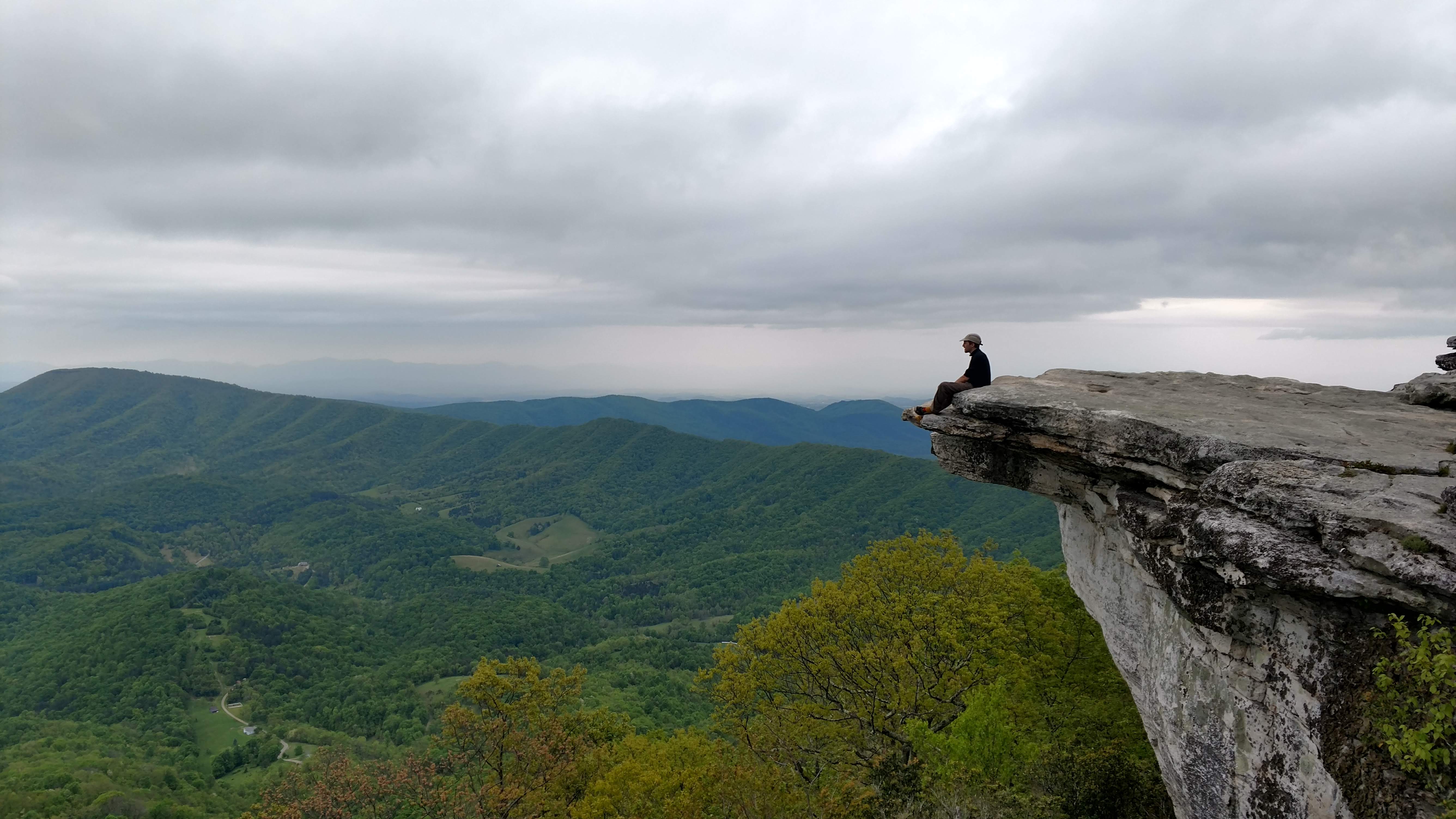 A hiker sitting on the exposed sandstone ledge at McAfee Knob, Virginia, looking out over rolling forested ridgelines receding into the distance under an overcast sky